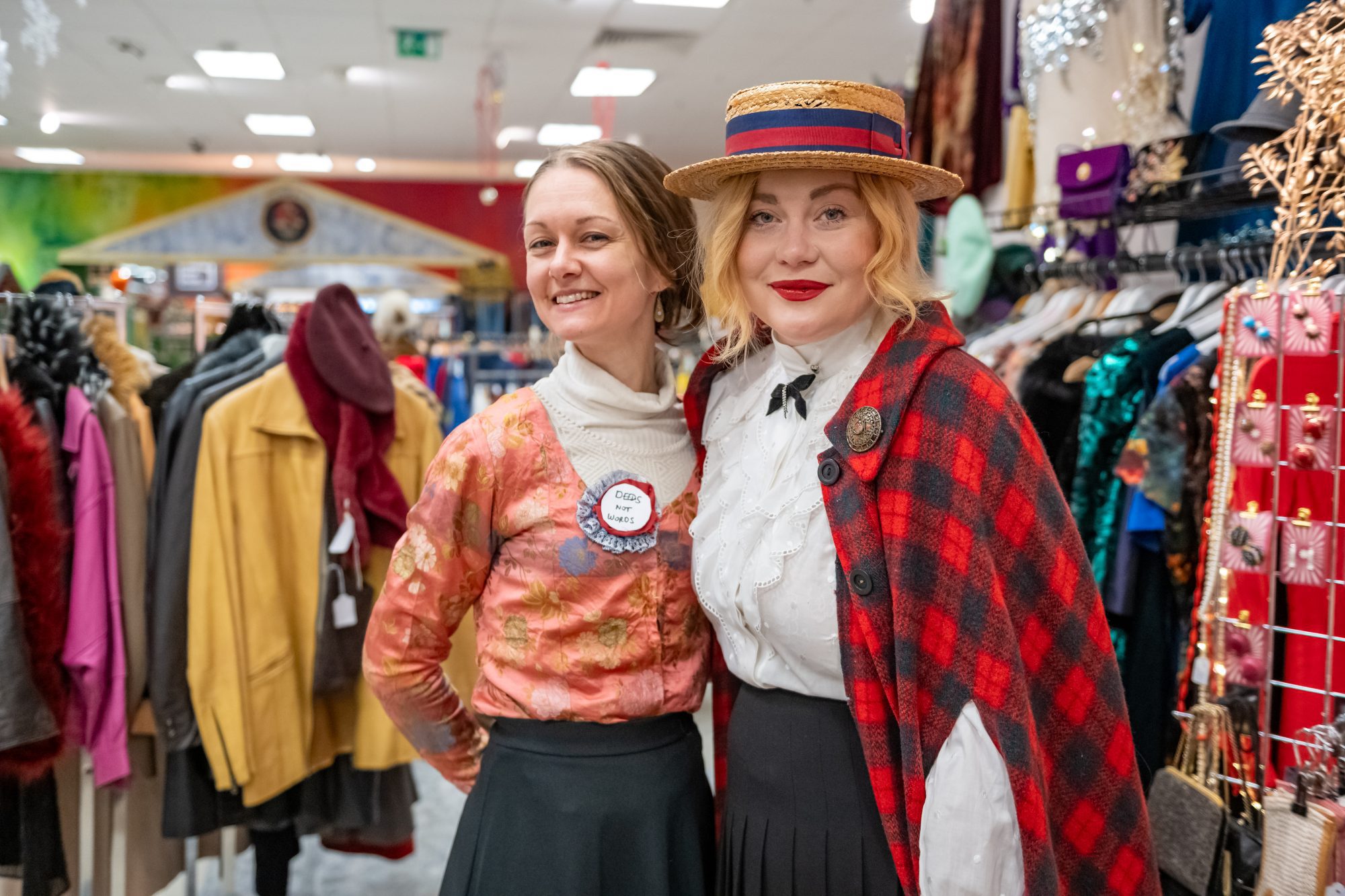 A woman in a red checkered cape and hat hugs another women in a clothing area at Sparks